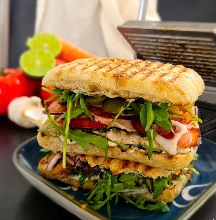 A gourmet panini filled with smoked chicken, premium prosciutto, dairy-free cheese, and tangy pickles, topped with peppery arugula and fresh tomatoes on a rustic surface.
